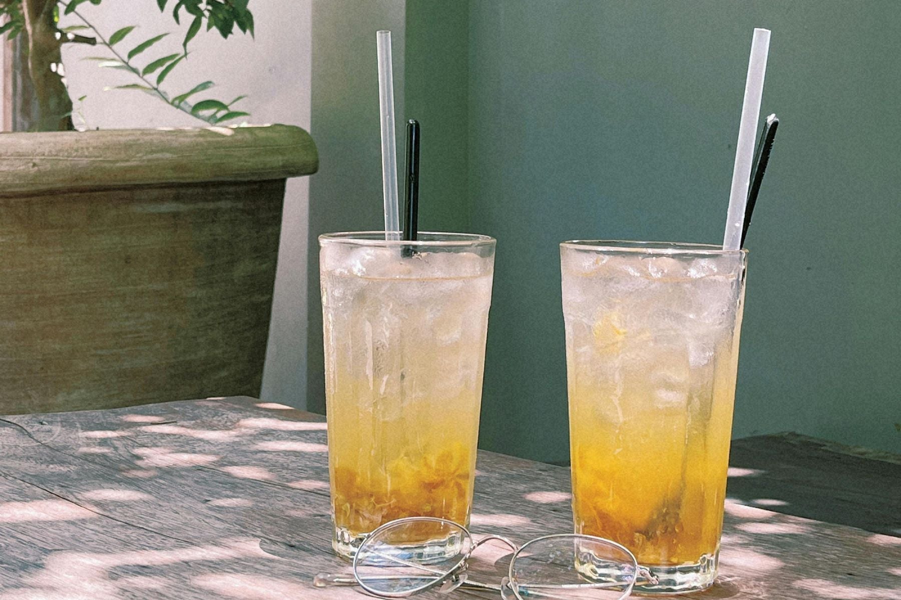 Two tall glasses of iced tea with straws on a sunlit outdoor table beside sunglasses and a potted plant