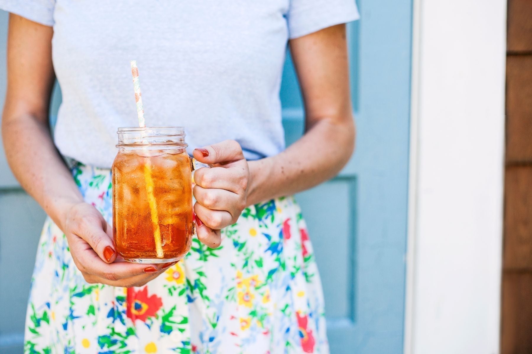 Person holding a mason jar of iced sweet tea with a straw, wearing a floral skirt outdoors by a blue door