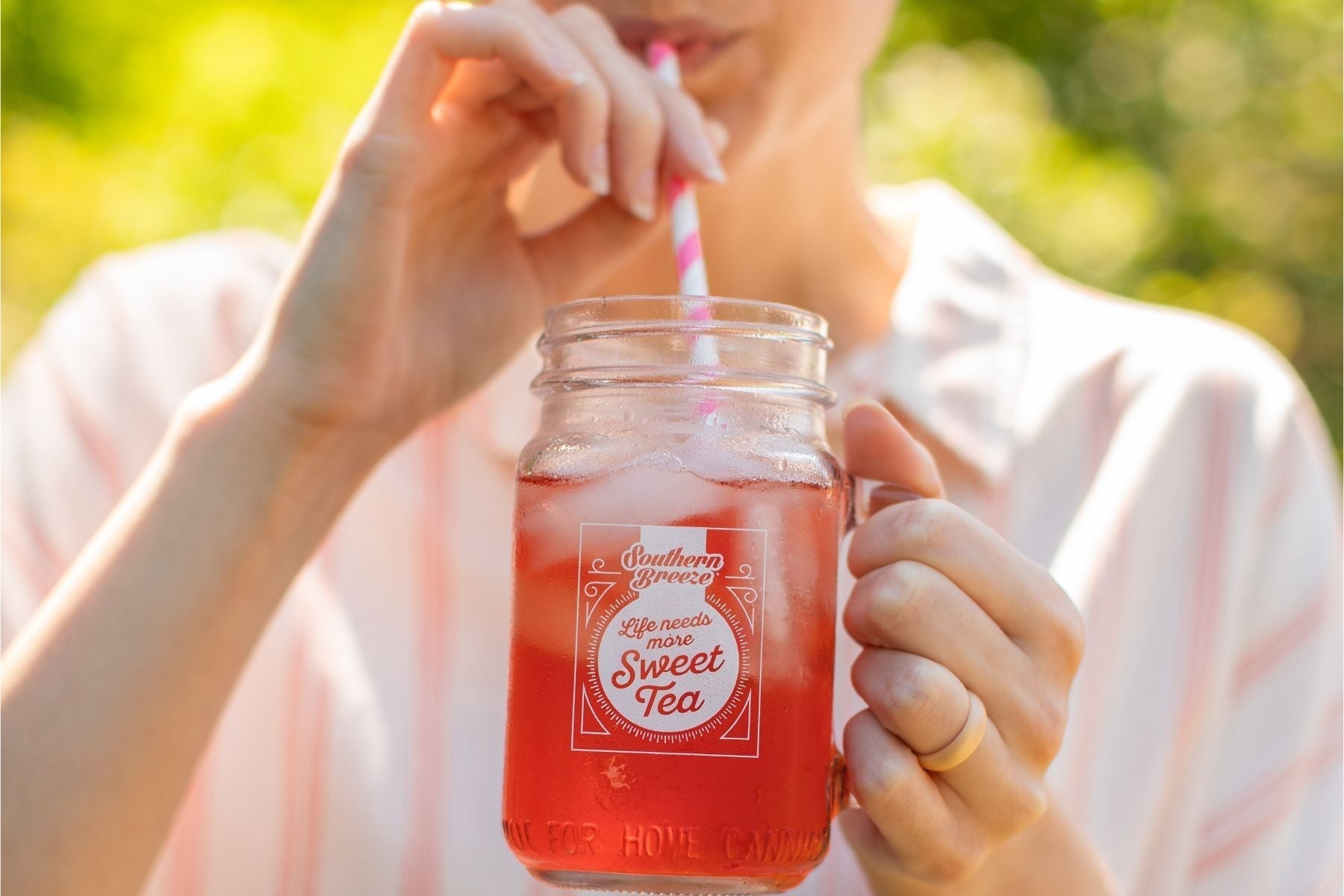 Person sipping iced sweet tea from a mason jar with a striped straw, featuring Southern Breeze branding, outdoors in warm sunlight.