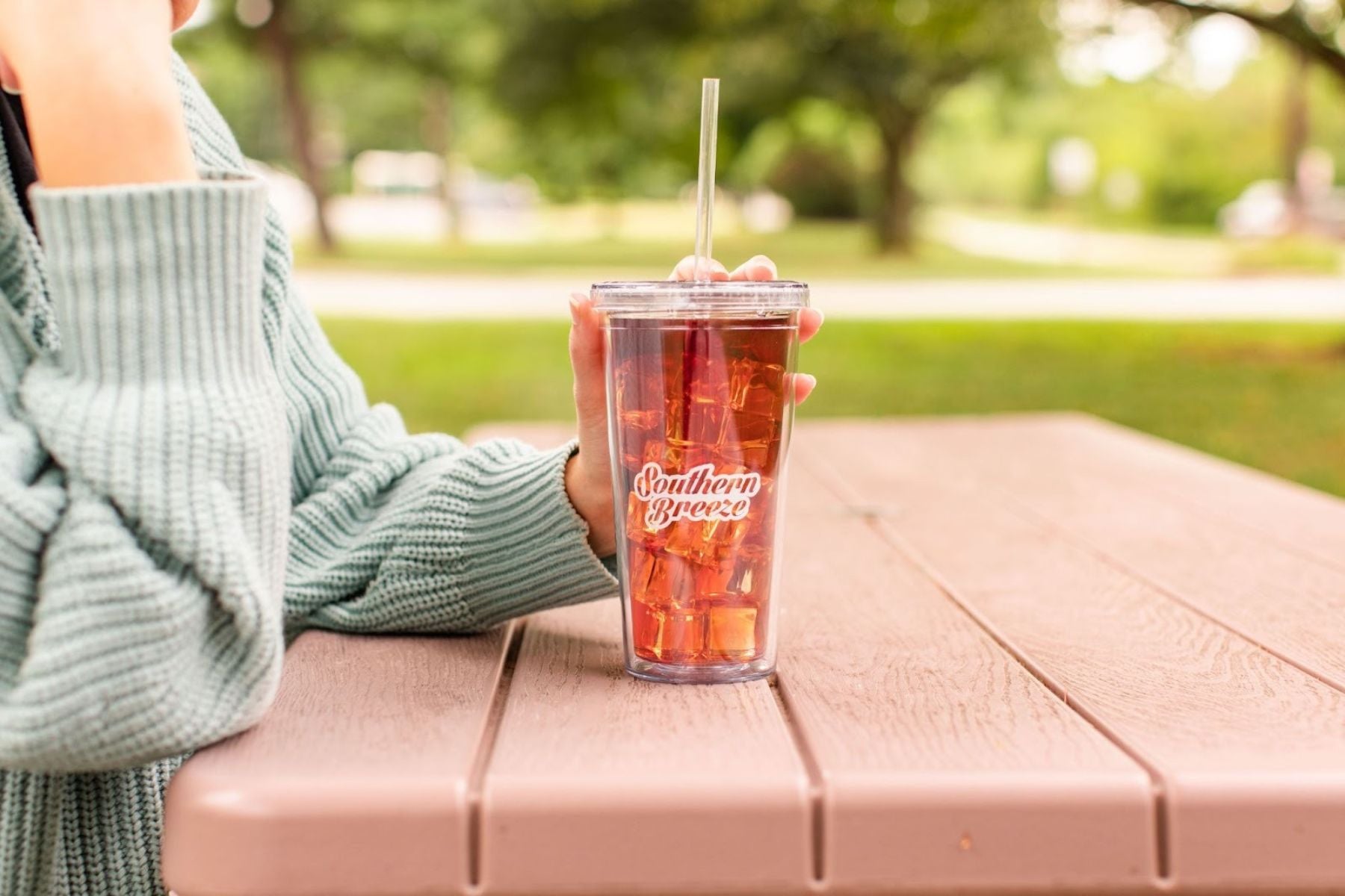 Person holding a clear tumbler of iced tea with a straw on an outdoor table