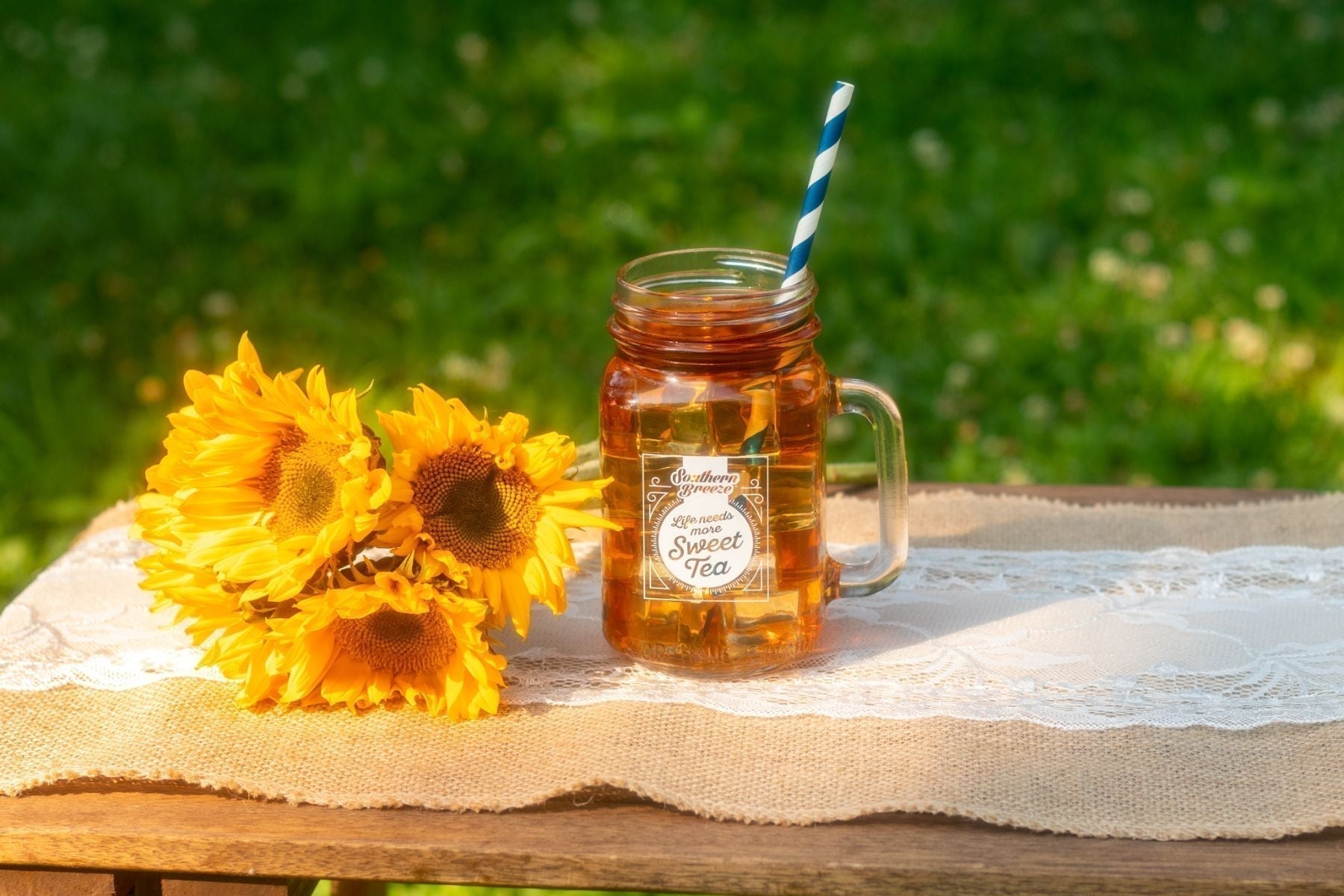Mason jar of iced sweet tea with a striped straw beside bright sunflowers on a rustic outdoor table.