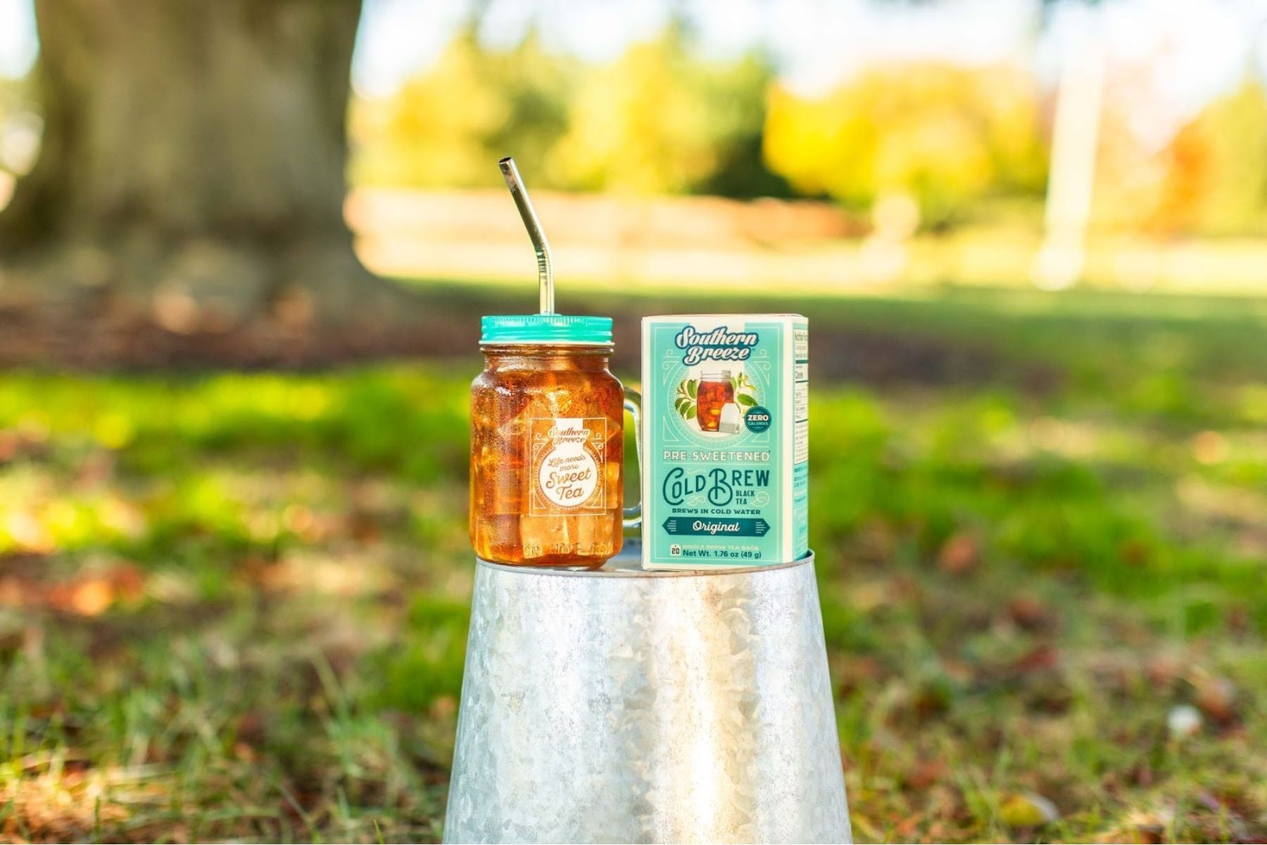 Mason jar of iced sweet tea with a metal straw beside a Southern Breeze cold brew tea box, placed outdoors on a metal stand with greenery in the background