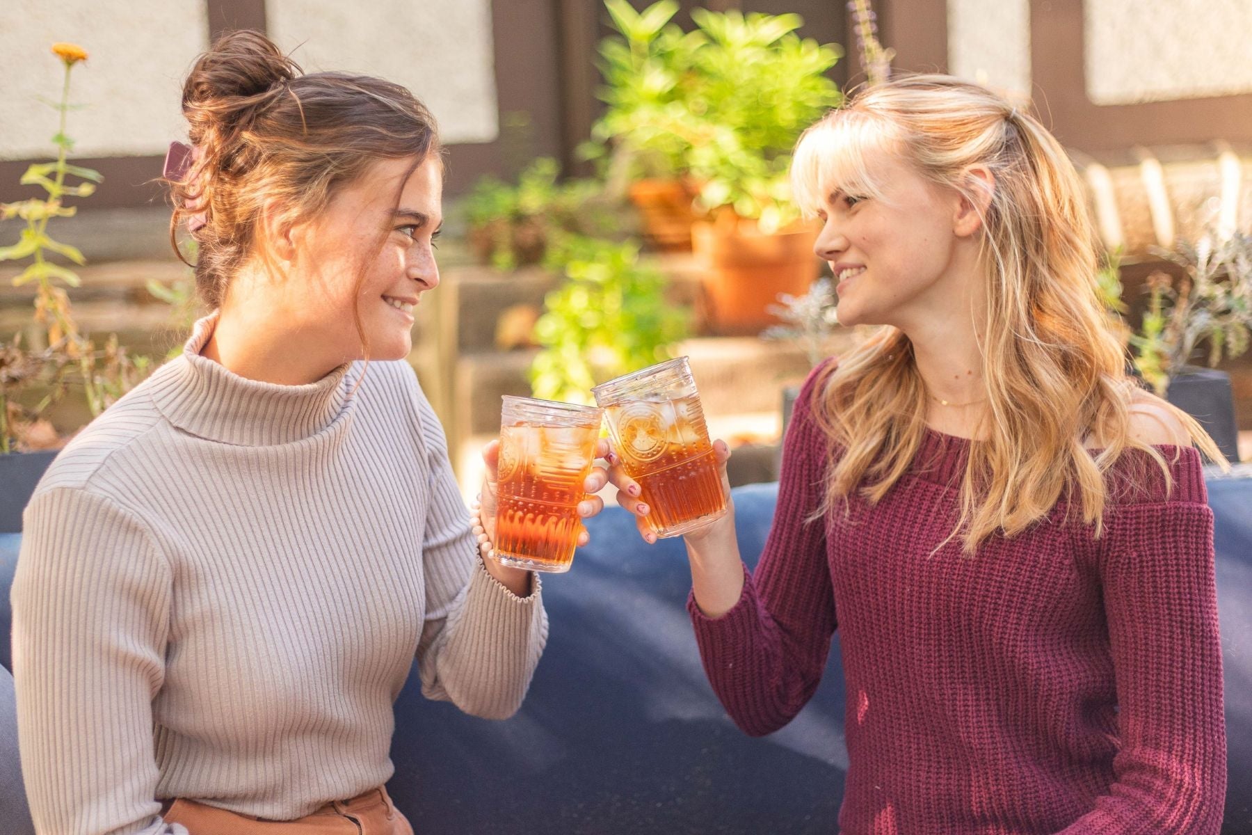 Two women smiling and holding iced tea glasses while sitting together outdoors in sunlight