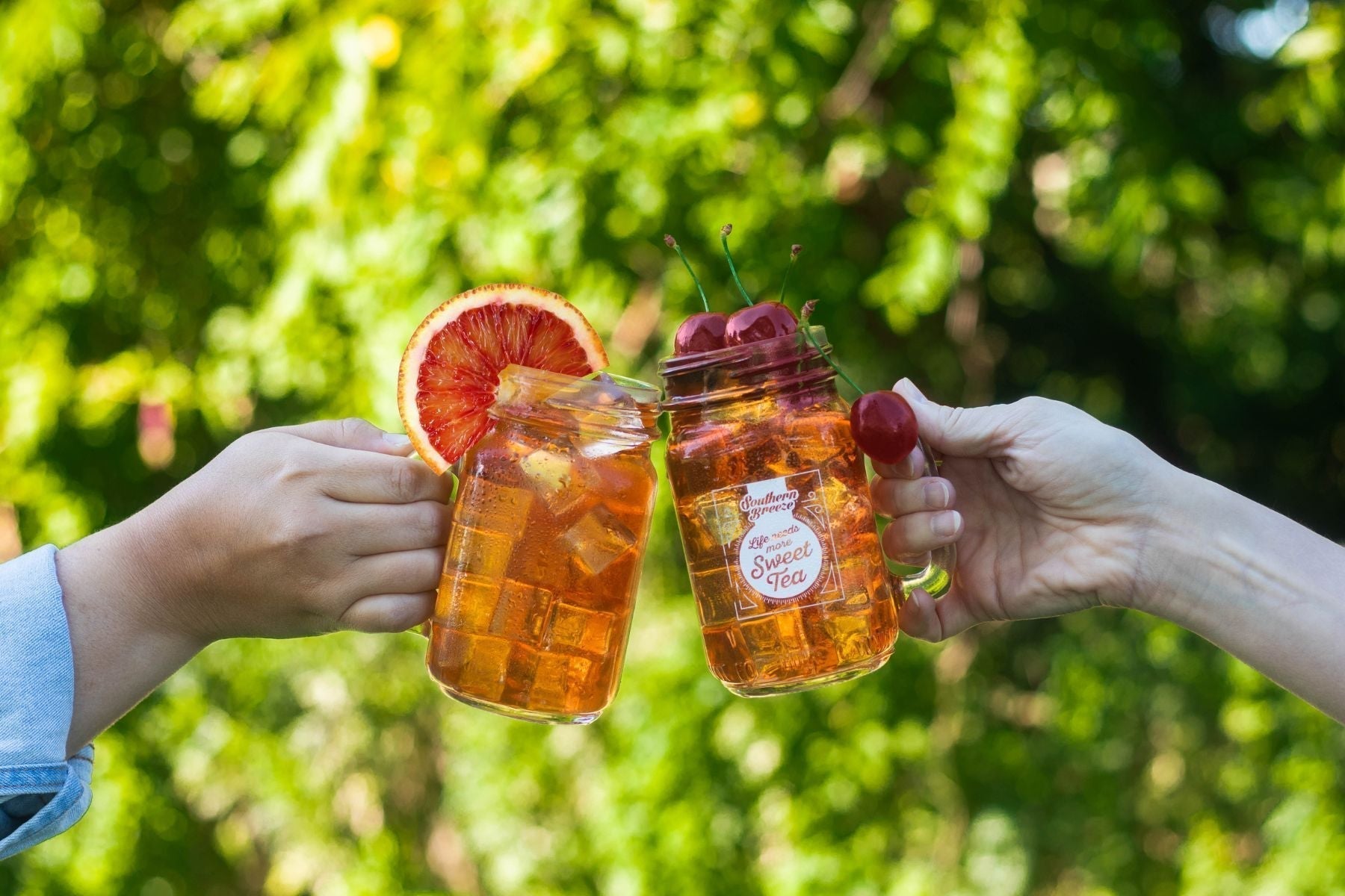 Friends holding mason jars of iced sweet tea garnished with cherries and citrus outdoors in bright summer sunlight