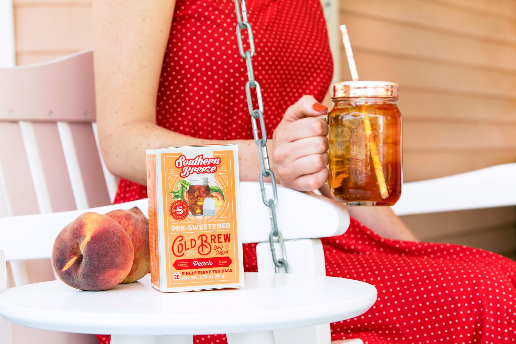 Woman in a red polka dot dress holding a mason jar of peach iced tea beside Southern Breeze Peach Cold Brew tea and fresh peaches on a porch swing table