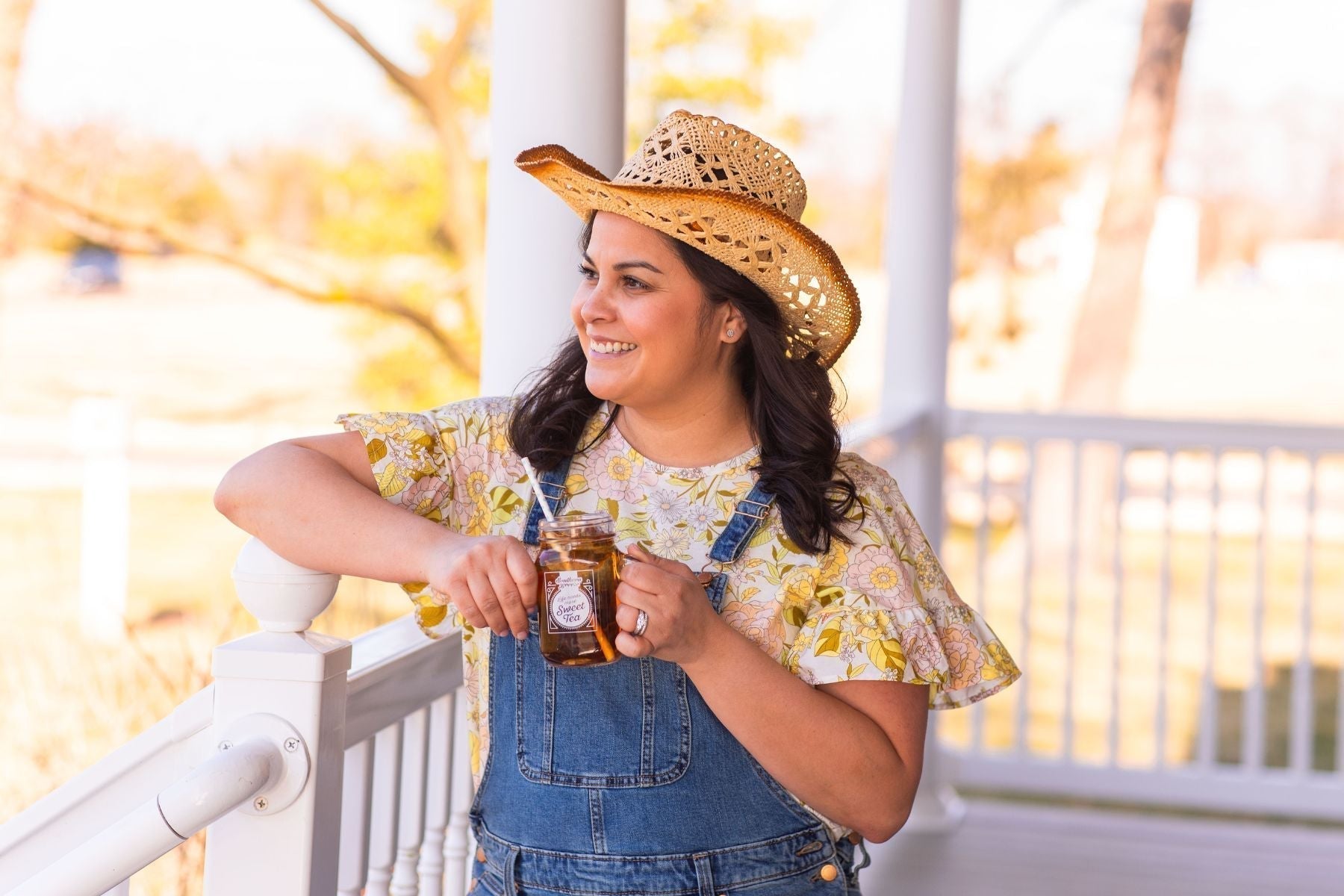 Woman wearing a hat and denim overalls holding a mason jar of Southern Breeze Sweet Iced Tea Original while standing on a porch