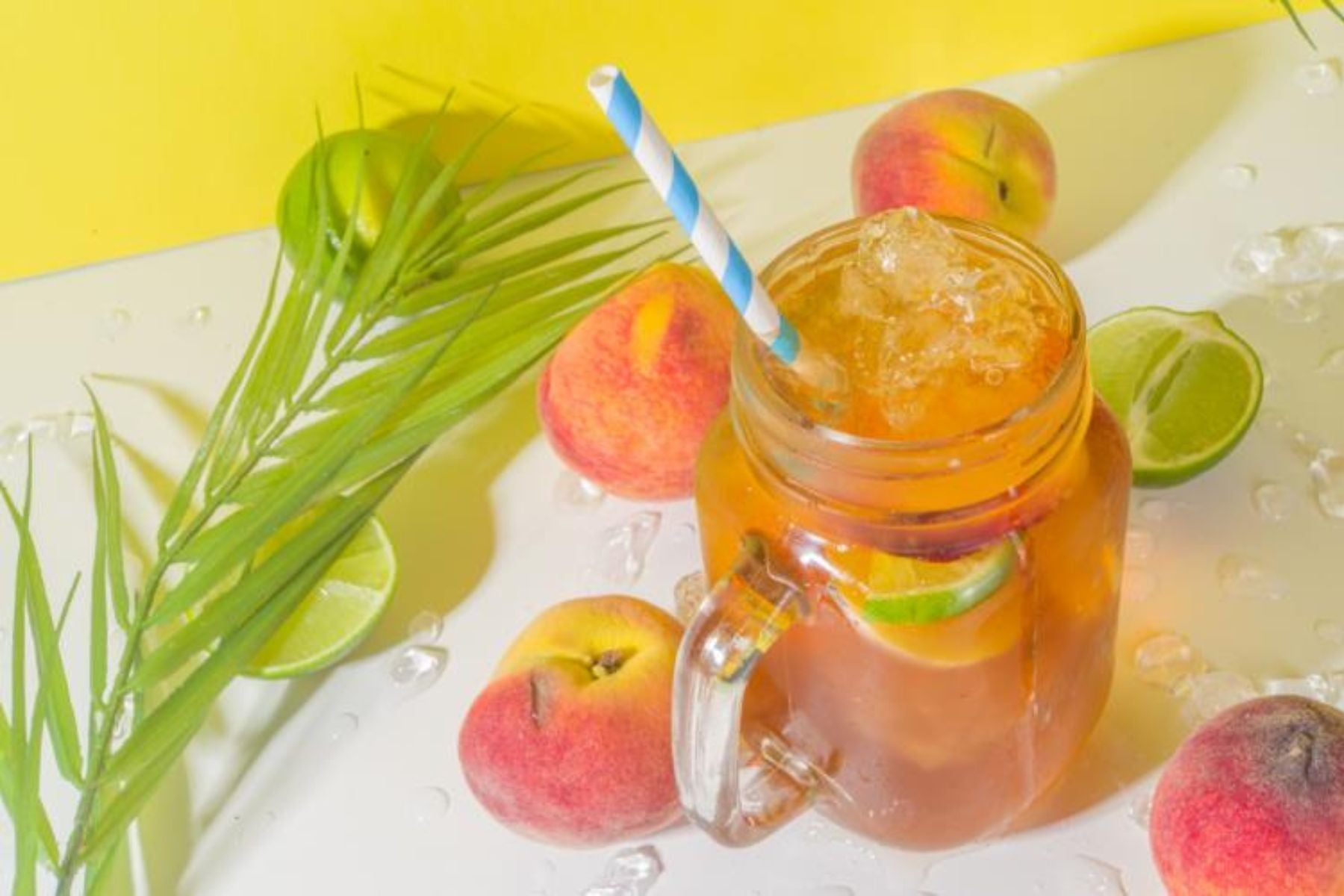Iced peach tea with lime slices and ice in a mason jar, surrounded by fresh peaches and limes on a bright background.