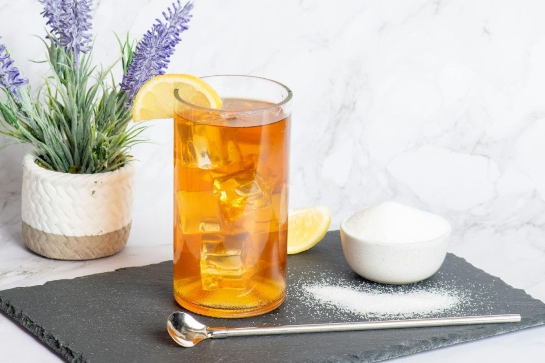Tall glass of iced tea with lemon and a bowl of sugar on a slate coaster.