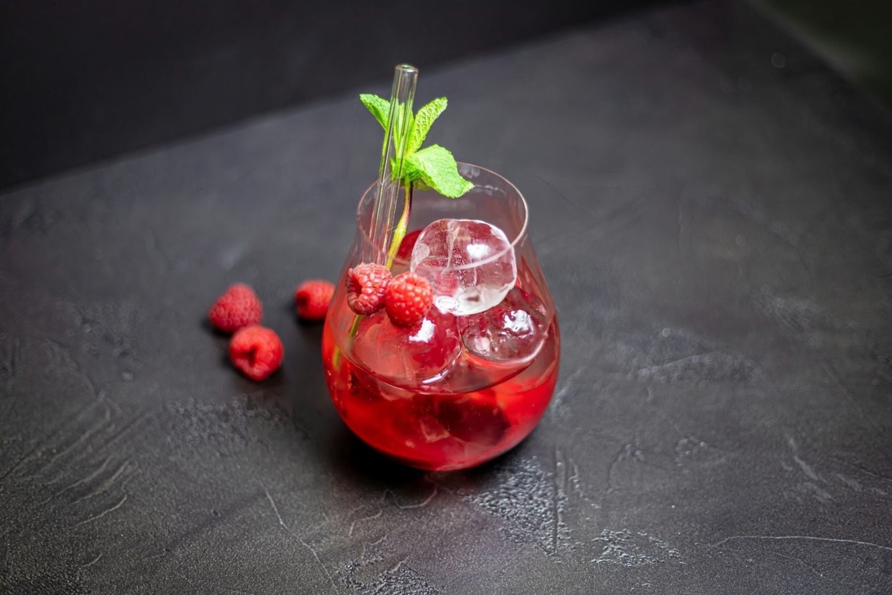 Iced raspberry drink garnished with mint and fresh berries on a dark surface.