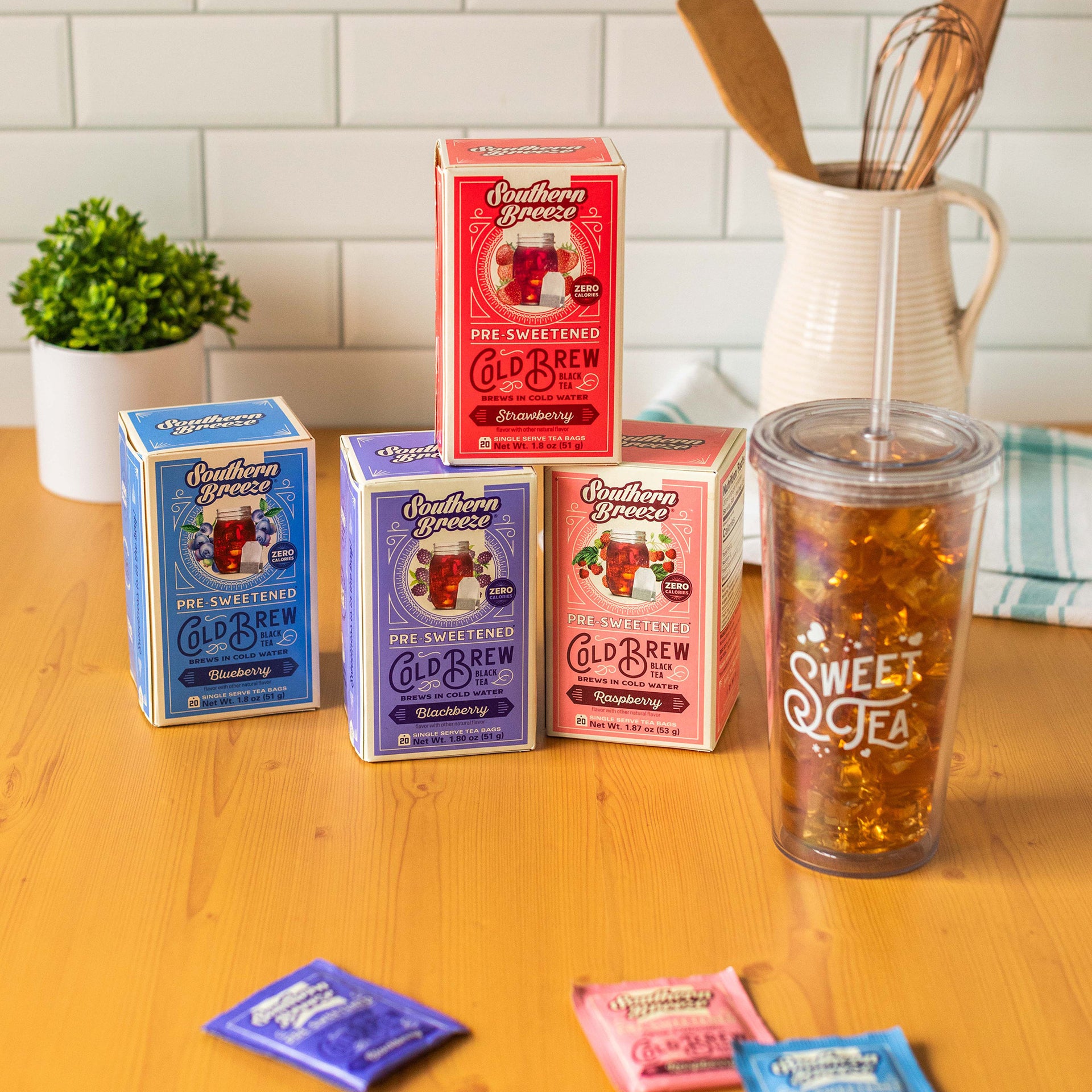 Four Southern Breeze Cold Brew tea boxes on a wooden countertop in a kitchen setting. The boxes are pink, blue, purple, and red. Flavors are Raspberry, Blueberry, Blackberry, and Strawberry. Next to them is a tumbler full of ice and sweet iced tea.