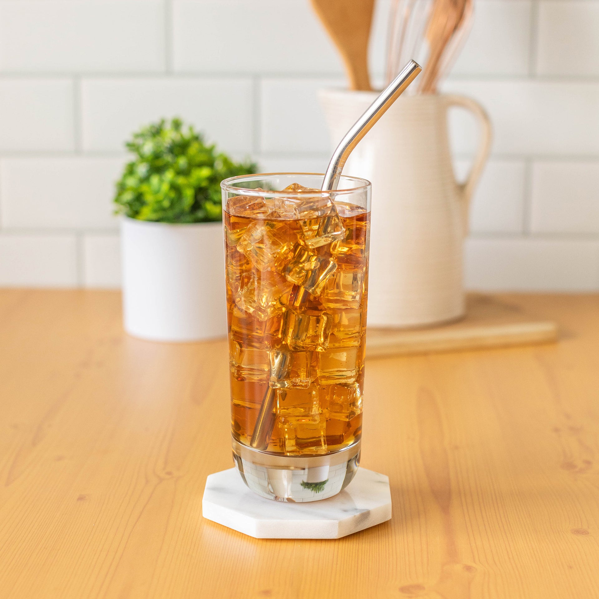 A clear glass tumbler filled with iced tea and ice, with a curved metal straw, resting on a small white coaster on a wooden table. A white pitcher and small plant are blurred in the background.