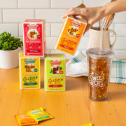 The boxes are in Guava, Mango, Tropical, and Watermelon flavors, next to a clear plastic cup with a lid that says "Sweet Tea" and is full of sweet tea and ice. A hand is holding the Mango box, and in the background are a plant and a kitchen tea towel on a wooden counter with a white backsplash.