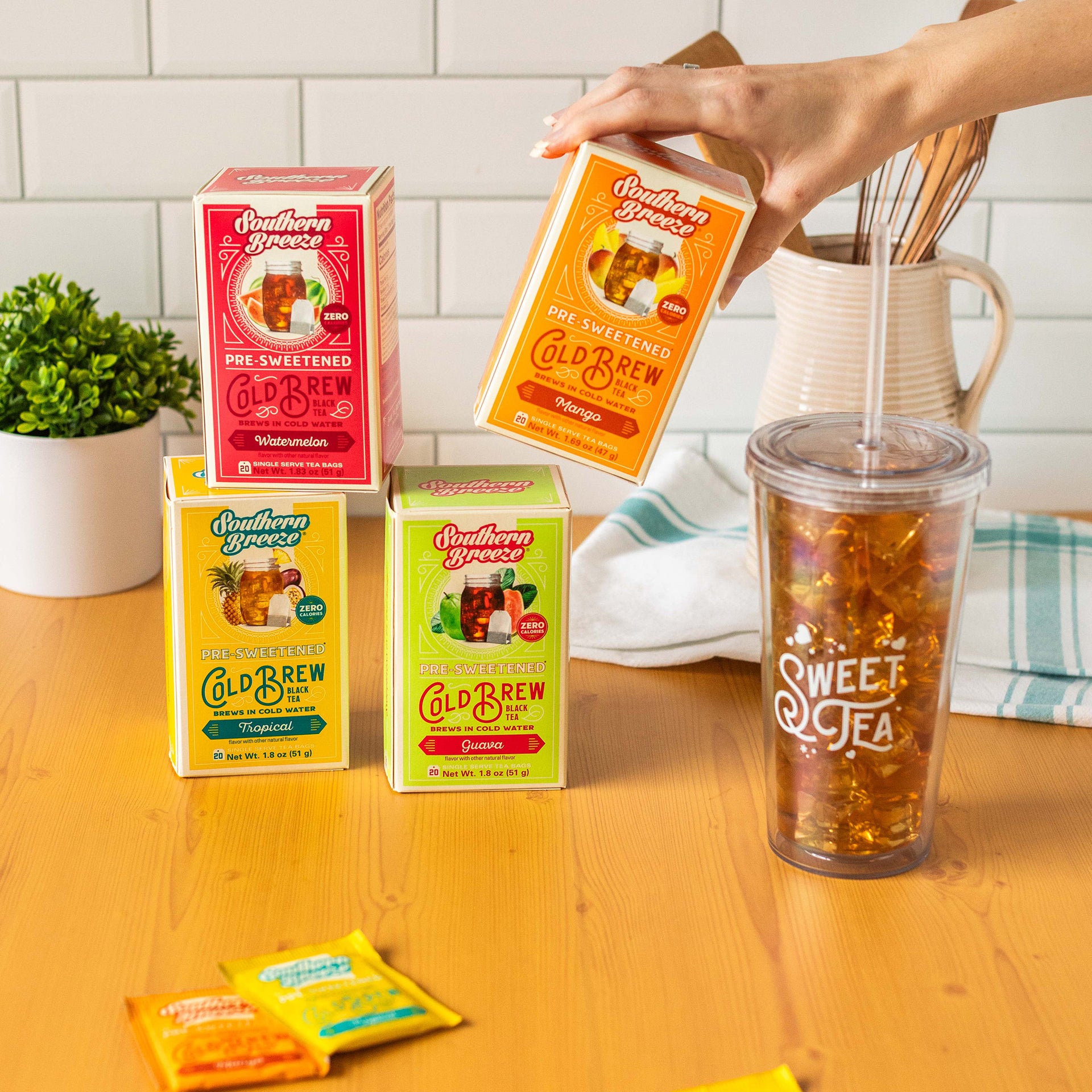 The boxes are in Guava, Mango, Tropical, and Watermelon flavors, next to a clear plastic cup with a lid that says "Sweet Tea" and is full of sweet tea and ice. A hand is holding the Mango box, and in the background are a plant and a kitchen tea towel on a wooden counter with a white backsplash.